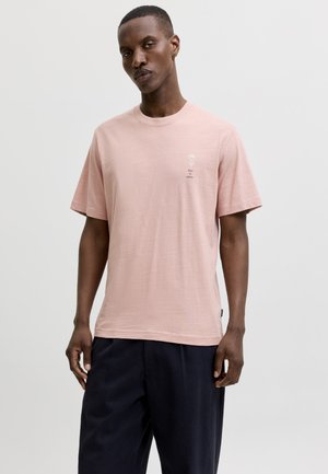 Homme portant un t-shirt rose clair à manches courtes avec un petit texte blanc sur la poitrine et un pantalon foncé et ample, sur un fond uni.