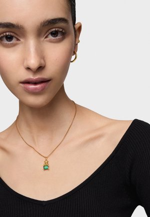 Złoty naszyjnik łańcuszkowy z zawieszką w kształcie zielonego kamienia, z wyróżniającym się teksturowanym wzorem i delikatnymi ogniwami, noszony na czarnej topie z prążkami.