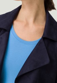 Lähikuva henkilöstä, jolla on päällään vaaleansininen pyöreäkauluksinen paita tummansinisän bleiserin alla, jossa on leveät läpät.