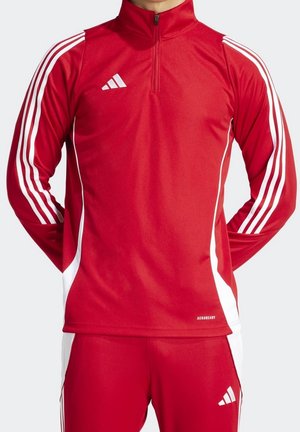 Homme portant une tenue de sport rouge Adidas avec des bandes blanches sur les manches et le pantalon, debout avec les mains dans le dos.
