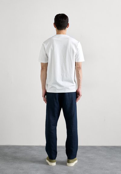 Hvid bomulds t-shirt med korte ærmer, parret med løse marineblå bukser. Modellen står på et gråt gulv og viser simple, rene linjer.