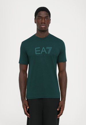 Man wearing dark green short-sleeve t-shirt with "EA7 Emporio Armani" logo, standing with arms relaxed against a plain white background.