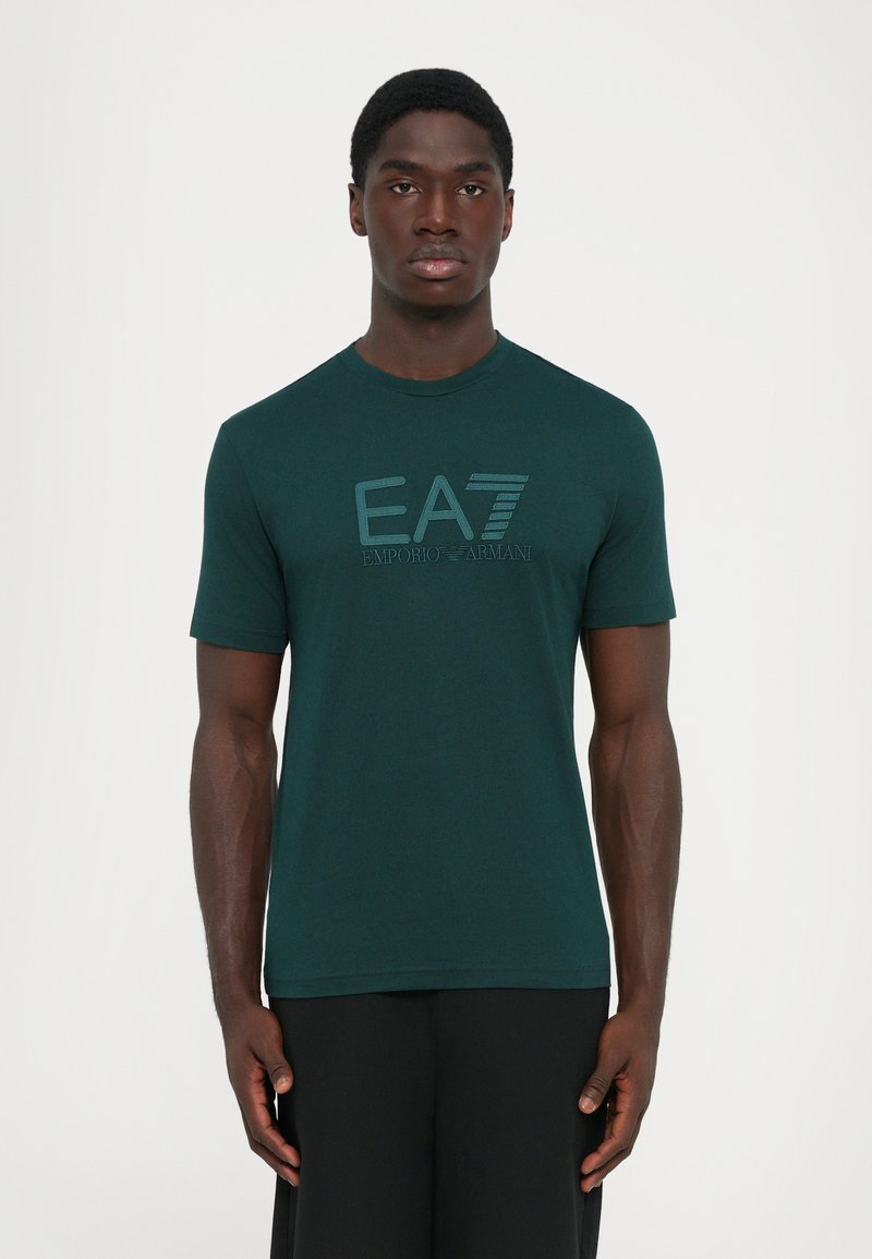 Vīrietis valkā tumši zaļu īso piedurkņu T-kreklu ar "EA7 Emporio Armani" logotipu, stāvot ar rokām atslābinātām pret vienkāršu baltu fonu.
