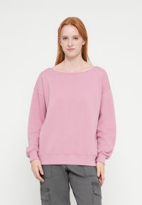 Világos rózsaszín pulóver széles nyakkal, hosszú ujjakkal és bordázott mandzsettával. Az anyaga puha, laza szabású, minimális dizájnelemekkel.