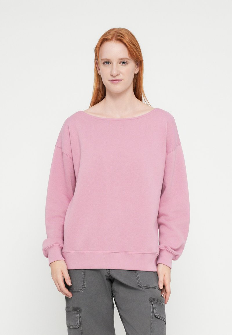 Világos rózsaszín pulóver széles nyakkal, hosszú ujjakkal és bordázott mandzsettával. Az anyaga puha, laza szabású, minimális dizájnelemekkel.