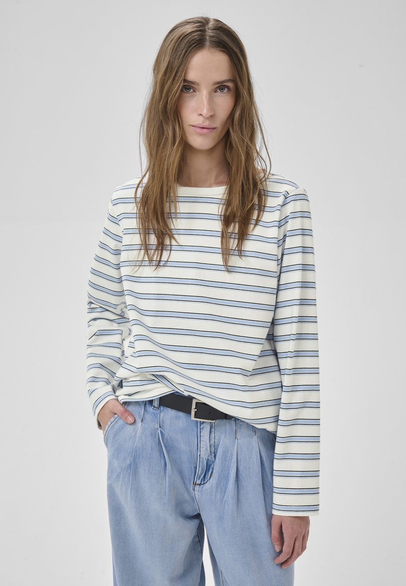 Mladá žena s dlhými hnedomi vlasmi, oblečená v bielej a modrej pruhovanej dlhorukávovej košeli a svetlomodrých vysokých rifliach s čiernym opaskom.