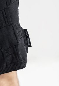 Maglione nero lavorato a maglia con motivi testurizzati, polsini a coste e un'etichetta del marchio visibile con lettering bianco su un lato.