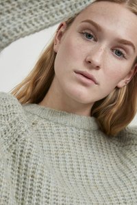 Hellgrüner Strickpullover mit strukturiertem Muster, mit einem gerippten Rundhalsausschnitt und überschnittenen Schultern. Nahaufnahme eines Gesichts.