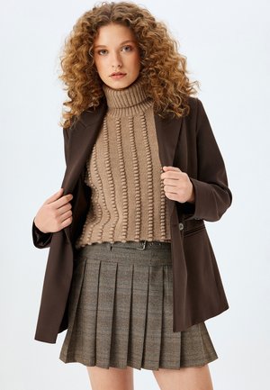 Blazer marron sur un pull à col roulé beige côtelé et une jupe plissée marron à carreaux. Les caractéristiques clés incluent la texture et les couleurs contrastées.