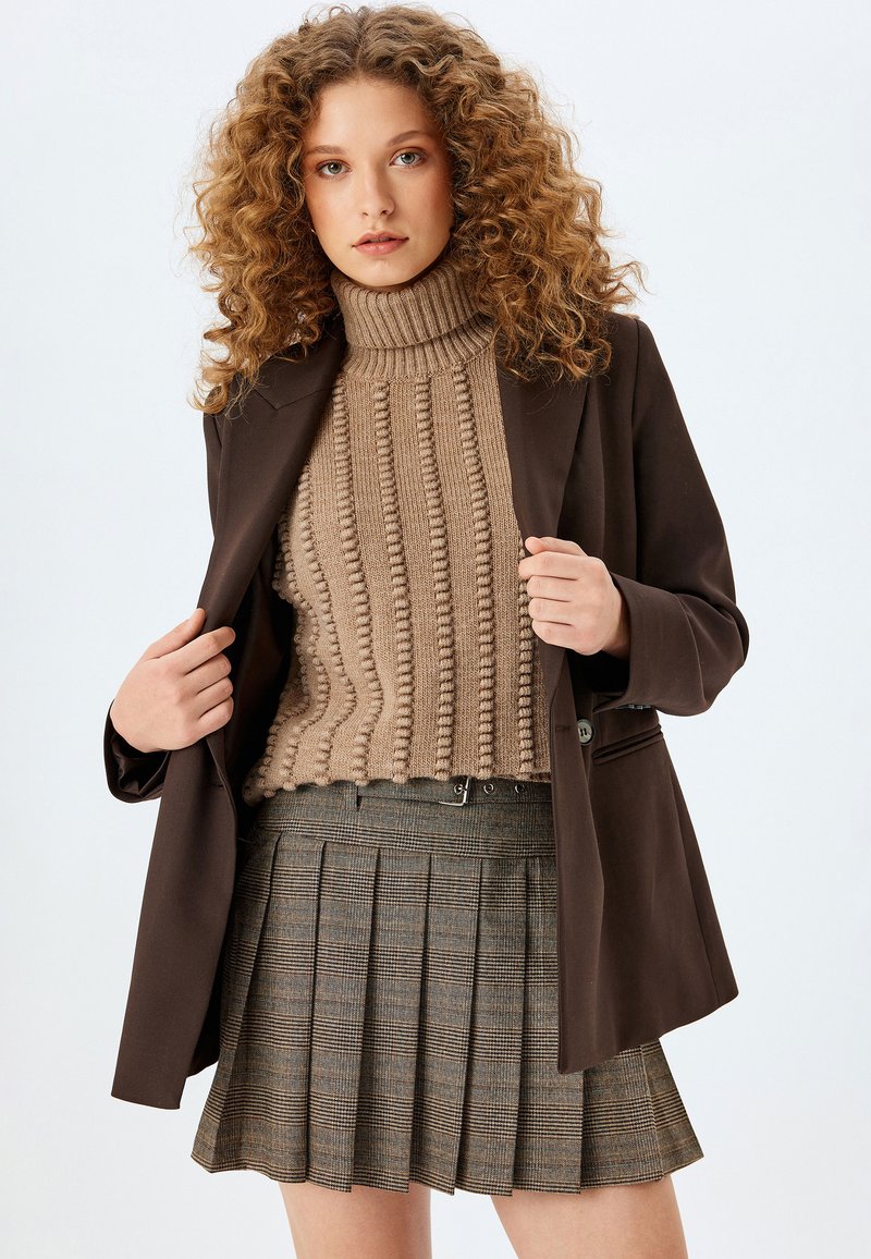 Blazer marron sur un pull à col roulé beige côtelé et une jupe plissée marron à carreaux. Les caractéristiques clés incluent la texture et les couleurs contrastées.