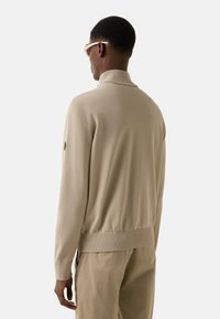 Beige knit sweater with a high collar and ribbed hem, featuring a black emblem on the left sleeve, worn over light-colored trousers.