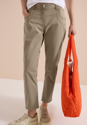 Kvinde iført khakibukser og beige sneakers, der holder en stor orange quiltet taske i højre hånd, venstre hånd i lommen.