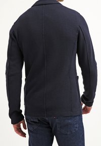 Giacca testurizzata blu navy con colletto su misura, cuciture posteriori e tasche laterali. Maniche lunghe e silhouette aderente.