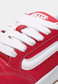 Adidași din nubuc roșu, cu șireturi albe, având un logo Vans pe limbă, accente albe pe laterale și detalii de cusătură vizibile.