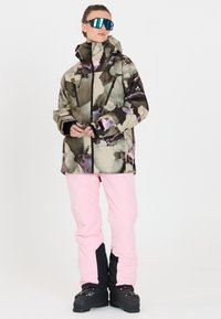 Schneejacke in einem mehrfarbigen Camouflage-Muster, mit hohem Kragen, Kapuze und schwarzem Reißverschluss, kombiniert mit hellpinkfarbigen Hosen und schwarzen Stiefeln.