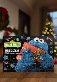 Socken-Geschenkset für Herren von Sesame Street in einer schwarzen Box, die ein pelziges blaues Monster zeigt, das ein rotes Geschenk mit goldenen Akzenten hält. Größe 40-45.