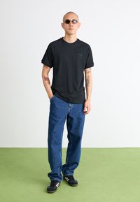 Czarny t-shirt z krótkim rękawem i ściślejszym kołnierzykiem, zestawiony z luźnymi niebieskimi dżinsami. Czarne sneakersy z białymi paskami i okrągłe okulary przeciwsłoneczne.