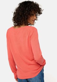 Pull en maille corail avec un col rond et des manches longues, présentant une texture douce et une coupe décontractée. Un jean en denim bleu complète la tenue.