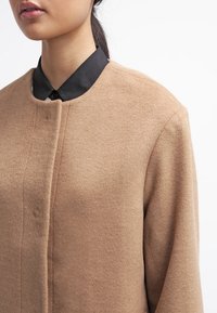 Płaszcz z wełnianego materiału w kolorze camel z okrągłym kołnierzem, zapięciem na pojedynczy guzik oraz fakturowanym materiałem. Pod nim widoczna jest czarna koszula z kołnierzykiem.