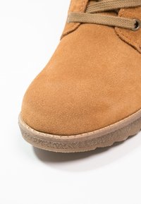 Gros plan sur l’orteil d’une botte en daim beige avec des lacets beiges et une semelle en caoutchouc texturée sur fond blanc.