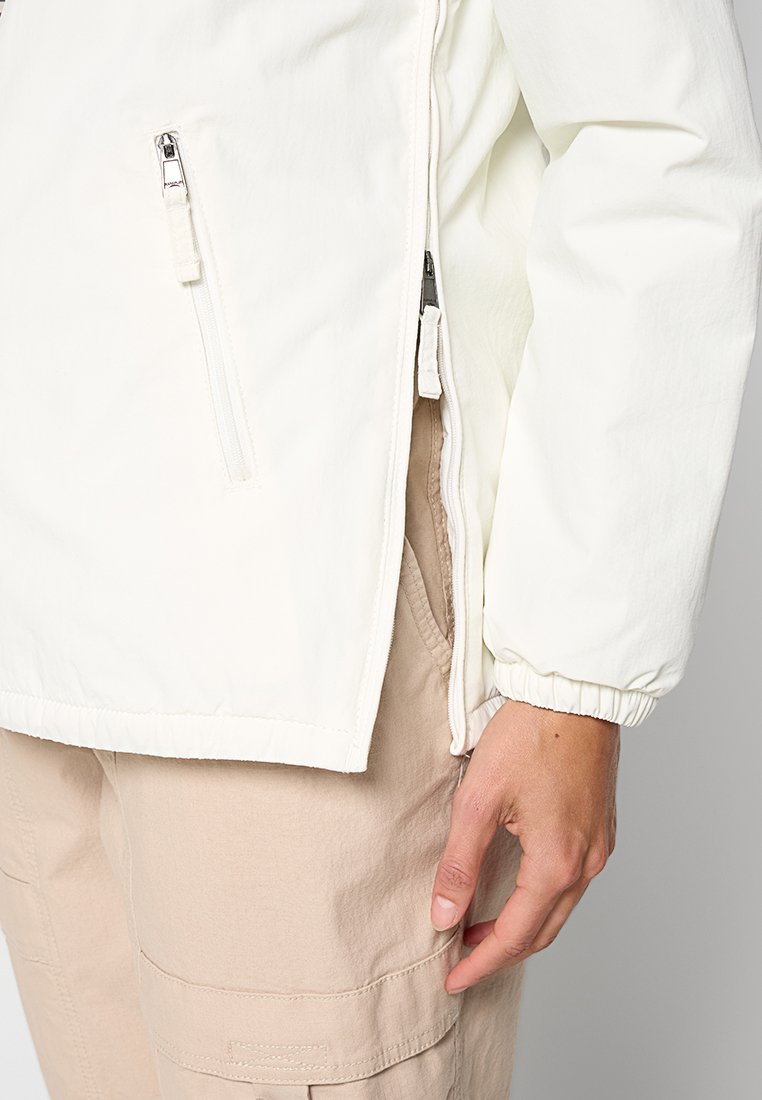 Nahaufnahme einer Person, die eine weiße Jacke mit Reißverschlusstaschen und beigen Cargo-Hosen trägt, mit der Hand in der Nähe einer Tasche.