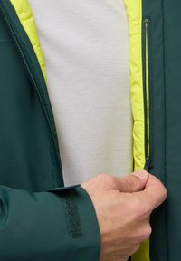 Veste extérieure verte avec une doublure jaune vif. Gros plan sur une main fermant la veste, mettant en valeur une sangle en velcro sur la manche.