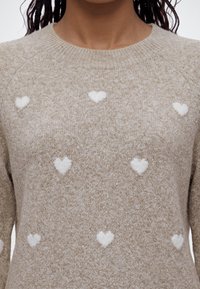 Pull beige tricoté avec des motifs de cœurs blancs porté par une personne aux cheveux bouclés foncés.
