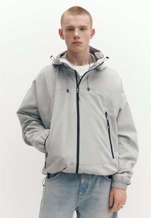 Mlad moški s kratkimi lasmi, oblečen v svetlo sivo kapuco z črnimi zadrgami in modre kavbojke, stoji z eno roko v žepu.