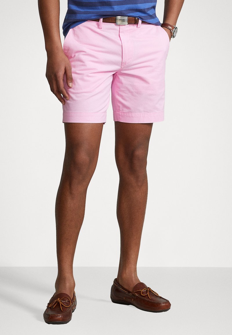 Roza bombažna kratka hlače z ravnim sprednjim delom, stranskimi žepi in zavihanimi robovi, obute z rjavimi usnjenimi čolnarskimi čevlji. Preprost, sproščen kroj.