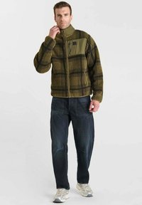 Fleecejack met een groen en zwart ruitpatroon, volledige rits en een borstzak. De jack is gecombineerd met donkerblauwe jeans en lichte sneakers.