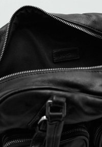 Interior de mochila de cuero negro con textura suave, con cierre de cremallera, forro de tela y una pequeña etiqueta. Accesorios incluyen cremallera plateada.