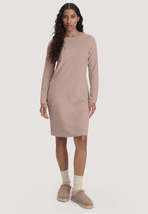 Frau mit langen lockigen Haaren, die ein einfaches beige knielanges Kleid mit langen Ärmeln trägt, mit cremefarbenen Socken und flauschigen Hausschuhen, steht vor einem einfarbigen Hintergrund.