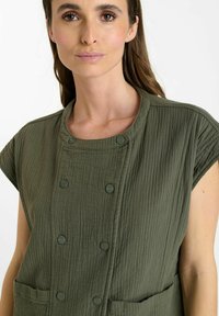 Tunic verde oliva con tessuto strutturato, caratterizzata da un neckline rotondo, maniche a cappuccio e due tasche frontali, chiuse con bottoni coordinati.