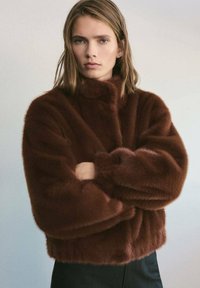 Giacca in pelliccia sintetica marrone con colletto alto, forma ampia e maniche voluminose. Texture morbida e senza chiusure visibili. Pantaloni neri sotto.