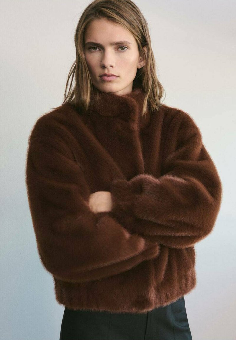 Giacca in pelliccia sintetica marrone con colletto alto, forma ampia e maniche voluminose. Texture morbida e senza chiusure visibili. Pantaloni neri sotto.