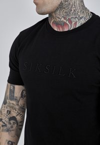 Melna pieguļoša T-krekls ar iespiestu "SIKSILK" logo priekšpusē. Materiāls izskatās mīksts, ar gludu tekstūru, īsām piedurknēm un apaļu izgriezumu.