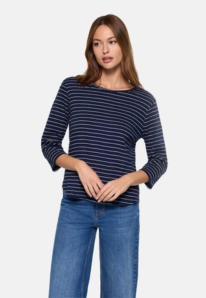 Mujer vistiendo una camiseta de manga larga azul marino con rayas horizontales blancas y jeans azules, de pie con las manos entrelazadas al frente.