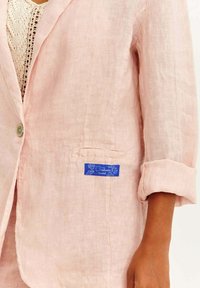 Světle růžový lněný blazer s jedním knoflíkem, ohrnovacími rukávy a modrou nášivkou na levé kapse. Měkká textura, ležérní design.