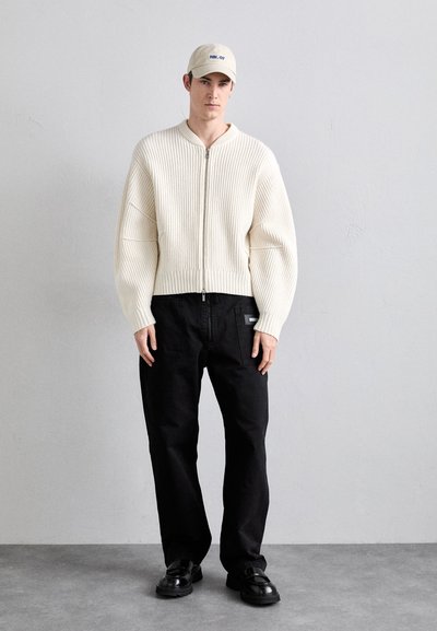 Chunky kremfarget ribbestrikket genser med glidelås foran, kombinert med svarte rette bukser og svarte sko, og med en beige caps.