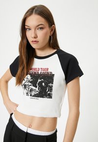 Geknipt t-shirt met een witte body en zwarte mouwen; beschikt over een grafische print met rode tekst en een zwart-wit afbeelding, ribbelstructuur.