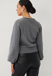 Grå strikket sweater med en t�ætsiddende kant og ballonærmer, der har en rund udskæring og perleaccentuer på ryggen. Sammen med kunstlæderbukser.
