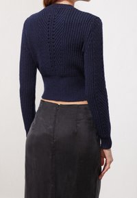 Pull en maille côtelée bleu marine à manches longues, avec une taille cintrée et un motif en losange dans le dos, associé à une jupe noire en tissu soyeux.