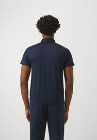 Mężczyzna z krótkimi, kręconymi włosami, ubrany w granatową koszulę polo z krótkim rękawem i dopasowane spodnie, stojący tyłem do gładkiej ściany.