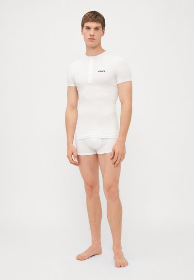 T-shirt bianco aderente con maniche corte e dettagli con bottoni; presenta un piccolo logo "VERSACE" nero sul petto. Indossato con biancheria intima bianca.