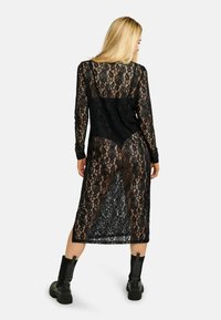 Fekete csipke hosszú ruha virágmintával, áttetsző textúrával és kontrasztos, egyszínű fekete hátrésszel. Térdig érő fekete csizmával viselve.