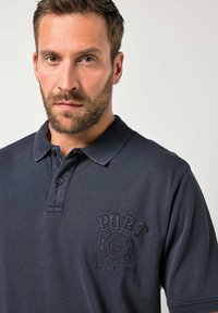 Námořnicky modré polo tričko z texturované bavlny, s límečkem a našitým logem "PORT" s kruhovým emblémem na levé straně hrudi.