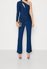 En axelbärande, utsvängd jumpsuit i metallisk blåt tyg med ett skärdetalj. Modellen håller en metallisk kuvertväska och bär lavendelfärgade klackar.