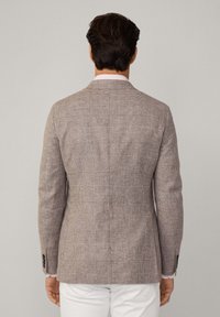 Blazer à carreaux marron avec un tissu texturé, coupe ajustée, revers crantés et accents en boutons noirs, porté sur un pantalon blanc.
