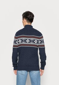 Mörkblå stickad tröja med hög krage, med ett festligt mönster av vita snöflingor, röda romber och zickzacklinjer.