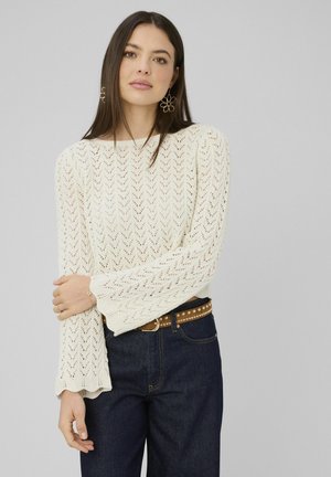 Jeune femme portant un pull à manches longues blanc à motifs, un jean taille haute foncé et une ceinture marron cloutée, debout les bras croisés.